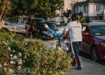 Volunteer assisting a person in a wheelchair on a city street surrounded by greenery and parked cars.