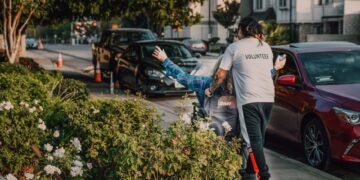 Volunteer assisting a person in a wheelchair on a city street surrounded by greenery and parked cars.